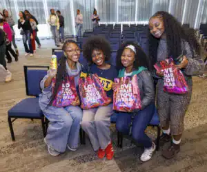 Four women sit together, smiling and holding colorful bags that read "LOVE HAIR YOU WEAR." One holds a can, and they are in a spacious venue with more people in the background.