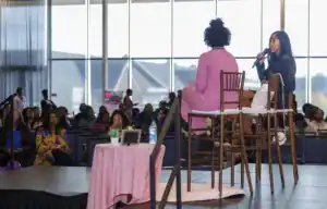 Two women sit on high stools at a panel discussion in front of an audience. One speaks into a microphone while the other listens. The venue has large windows and a decorated stage. Audience members are engaged and attentive.