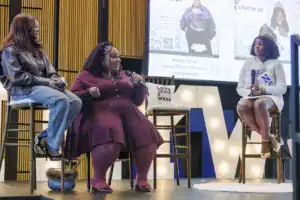Two women on stage during a panel discussion. One speaks into a microphone, wearing a maroon dress, while the other sits beside her, wearing a black jacket and jeans. A banner with event details is visible in the background.