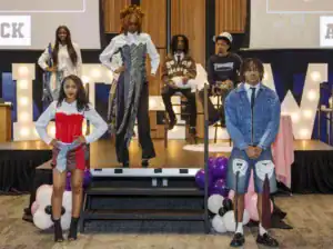 A fashion show featuring diverse models on a stage. Two models in the foreground showcase unique outfits made from ties and fabric, while others prepare in the background, highlighting creativity and style.