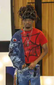 A model walks the runway wearing a red graffiti-style shirt and a distressed denim jacket, accessorized with layered necklaces and a unique spiked headpiece. The background features warm wooden panels.