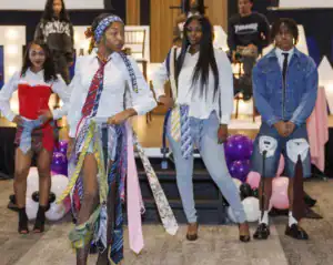 A group of four models showcases a fashion theme on a runway. They wear creative outfits made from ties, with one model in a corset and shorts, while another sports a denim jacket and shorts. Colorful balloons decorate the background.