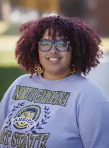 Smiling individual with curly, reddish hair and glasses, wearing a gray sweatshirt featuring "North Carolina A&T State." Background shows blurred greenery and buildings.
