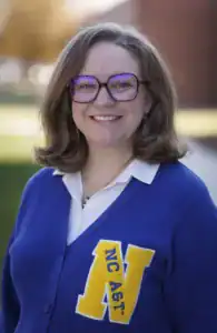 A woman with shoulder-length hair smiles, wearing large glasses and a blue cardigan featuring an "NC A&T" emblem. She stands outdoors with a blurred background of autumn foliage.