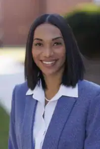 A woman smiles while wearing a blue blazer over a white blouse. She has straight, dark hair and is outdoors with a blurred background of greenery and brick walls.