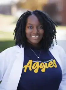 A woman with curly hair smiles while wearing a white lab coat over a dark blue t-shirt that says "Aggies" in yellow letters. She stands outdoors with a blurred background.