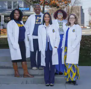 Five people in white lab coats stand on steps outside, smiling. They represent diverse backgrounds, wearing various styles underneath. The setting features a street sign and a blurred cityscape in the background.
