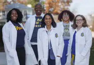Five women in white lab coats stand together outside, smiling. They are wearing shirts representing North Carolina A&T State University and showcasing school colors. The background features trees and blurred buildings.