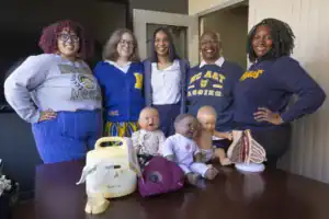 A group of five women stands by a table with educational dolls, a breast pump, and anatomical models. They are dressed in university sweatshirts, smiling at the camera in a well-lit office space.