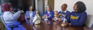 Five women sit around a table, smiling and engaging, with baby dolls and teaching tools in front of them. The setting suggests a classroom or training environment focused on childcare or healthcare.