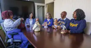 A group of five women sit around a conference table, smiling and interacting. They are holding dolls and teaching materials, suggesting a discussion on education or child care. A large screen is visible in the background.