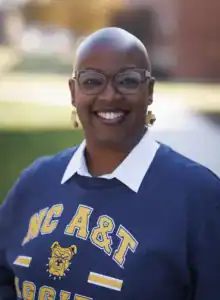 A smiling woman with a bald head wears glasses and a navy sweatshirt featuring "NC A&T" in yellow. She stands outdoors with a blurred background of grass and buildings.