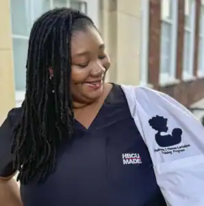 A smiling woman with long, dark hair in locs wears a black shirt with "HBCU MADE" printed on it. She holds a white lab coat embroidered with "Pathway 2 Human Lactation Training Program.