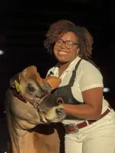 A smiling woman with curly hair poses with a brown cow, wearing a white shirt and overalls. The cow has a halter and ear tag. They are both in a well-lit farm setting.