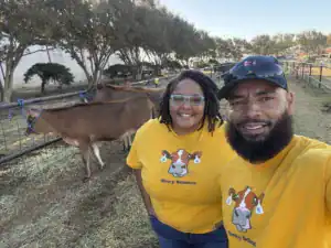 A man and a woman with glasses, both wearing yellow shirts featuring a cow's face, smile in front of two cows in a pen.Trees and a barn are visible in the background.