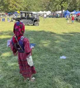A child in traditional attire holds a tablet, watching a utility vehicle in a grassy field filled with stalls and people at a fair. Colorful tents and chairs are visible in the background.
