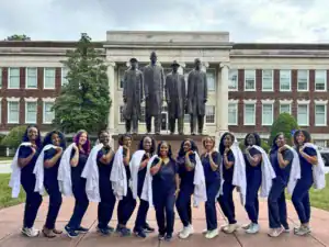 A group of women in navy scrubs and white lab coats pose confidently in front of a historic building and a statue. They smile and showcase their pride and achievement.