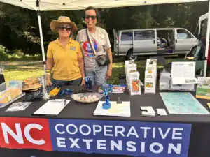 Two women stand behind a booth with various brochures and materials. One wears a yellow shirt and sun hat, while the other has glasses and a blue shirt. A gray van is parked in the background.