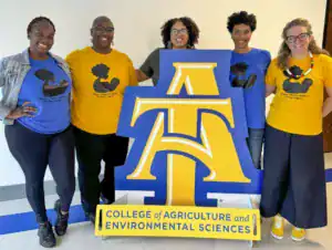 Five people stand together, wearing colorful t-shirts promoting the Pathway 2 Human Lactation Training Program, next to a large sign for the College of Agriculture and Environmental Sciences.
