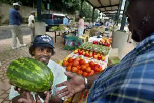 A vendor showcases a watermelon to a customer at a bustling farmers market filled with various fruits and vegetables, with other shoppers in the background.