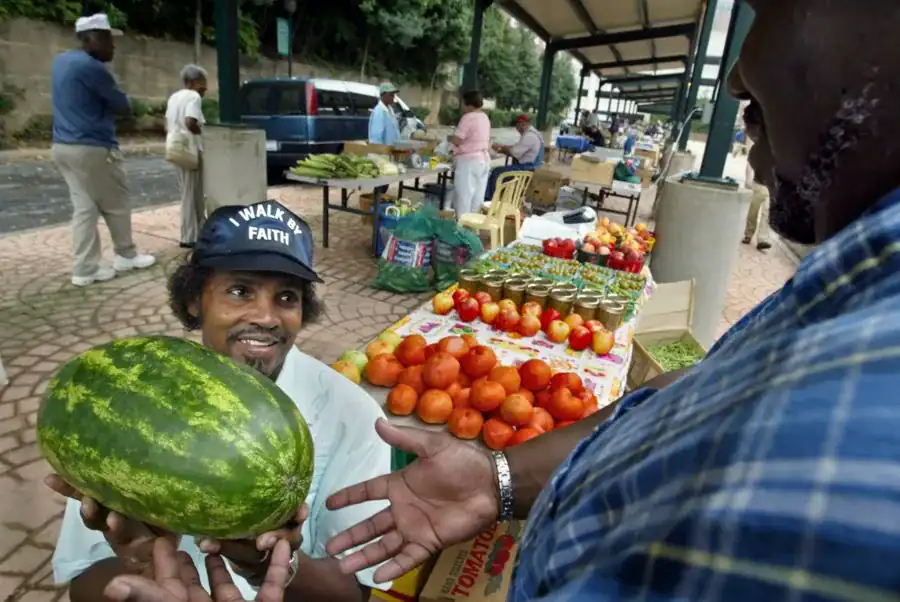 A vendor showcases a watermelon to a customer at a bustling farmers market filled with various fruits and vegetables, with other shoppers in the background.