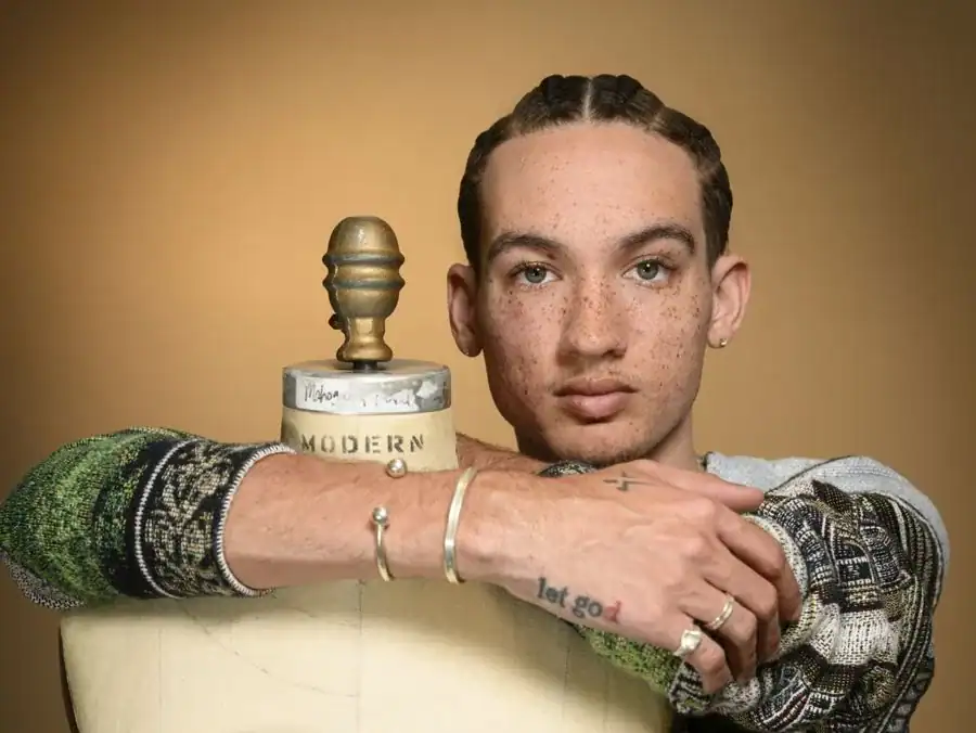 A young person with braided hair and freckles poses, resting their arms on a mannequin. They wear a colorful sweater and several rings, with a tattoo visible on one arm. The background is a warm, neutral tone.