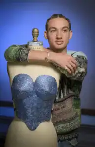 A person with braided hair leans against a dress form adorned with a shimmering blue top, wearing a colorful sweater and multiple rings, set against a blue background.