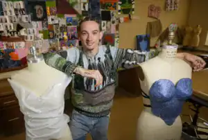 A person with braided hair poses between two dress forms, showcasing a white draped design and a blue embellished top. Behind them, a colorful collage of fashion sketches and designs decorates the wall.
