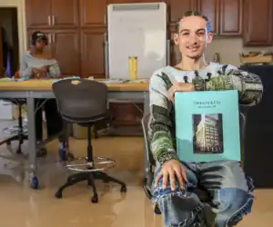 A young person with braided hair smiles while holding a large book titled "Tiffany & Co. THE LANDMARK." In the background, another individual appears focused on a work table.