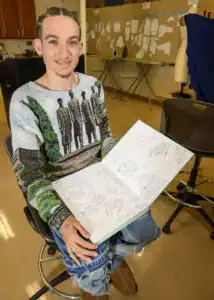 A young person with braided hair sits in a chair, holding an open notebook filled with handwritten notes and drawings. They wear a colorful sweater and distressed jeans, with their hand resting on the notebook.