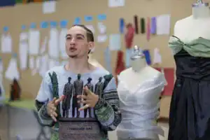 A person with light skin and braided hair gestures while speaking in a sewing studio, surrounded by garments displayed on mannequins and fabric samples on the wall.