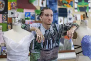 A young person with braided hair leans beside a dress form. They wear a colorful, patterned sweater, with a white garment draped on another nearby dress form. Art pieces decorate the background.