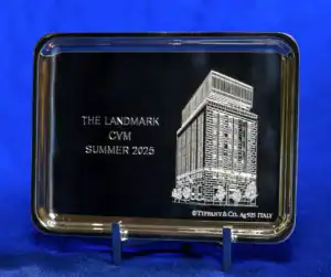 A silver plaque featuring an engraved building design, with the text "THE LANDMARK CVM SUMMER 2025" and "Tiffany & Co. Ag 925 ITALY" at the bottom. The background is a blue fabric.