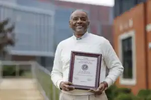 A smiling man holds a framed award outside a building, wearing a white sweater and blue shirt. The plaque recognizes his achievements in public service and agricultural leadership.