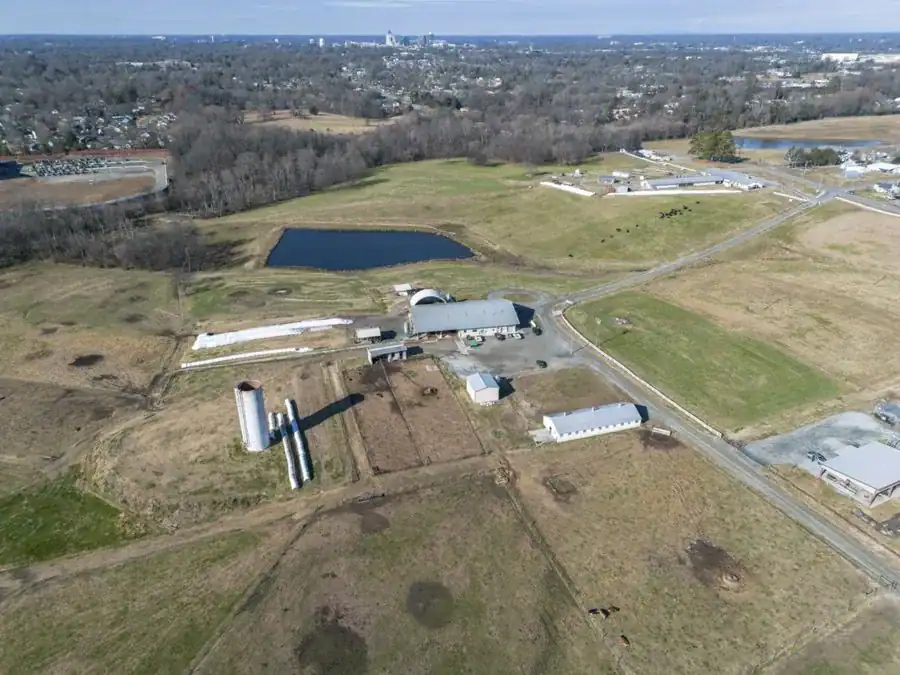 Aerial view of a rural landscape featuring several buildings, silos, and a pond, surrounded by fields and wooded areas. The skyline of a city is visible in the background.