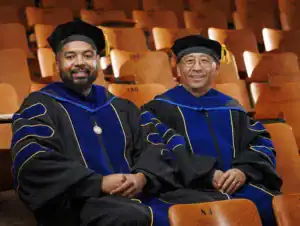 Two smiling men in academic regalia sitting on a wooden bench in a lecture hall. They wear black gowns with blue and gold accents, and caps with tassels. The background features empty wooden seats.