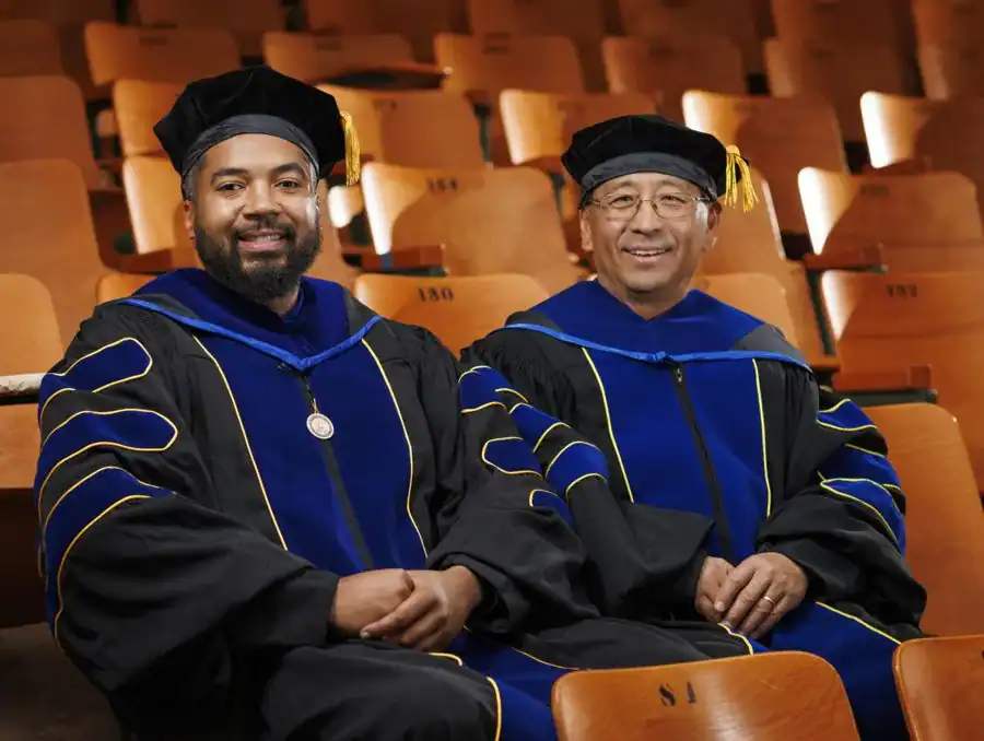 Two smiling men in academic regalia sitting on a wooden bench in a lecture hall. They wear black gowns with blue and gold accents, and caps with tassels. The background features empty wooden seats.