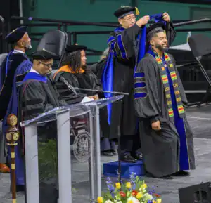 A graduate in a black gown and colorful sash receives their hood from a faculty member during a graduation ceremony. Other faculty members and guests are present. A podium with a seal is in the foreground, surrounded by flowers.