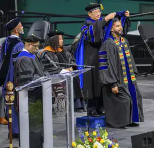 A graduate receives a hood from a faculty member during a commencement ceremony, while other faculty and attendees observe. Floral arrangements are visible in the foreground.