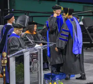 Graduates in academic regalia celebrate at a ceremony. One graduate smiles and fist pumps while receiving recognition. Others, including faculty, observe from the side. A podium with a microphone is visible in the foreground.