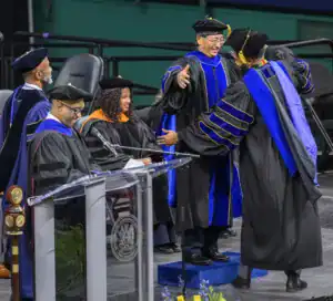 Graduates in academic regalia celebrate on stage during a commencement ceremony, with one receiving a citation. Faculty members observe, and a podium with a microphone is in the foreground.