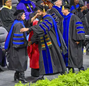 Graduates in academic regalia celebrate at a commencement ceremony. One graduate receives congratulations from a faculty member, while others in the background observe the moment.