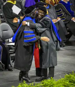 Two graduates in academic regalia embrace joyfully on stage, celebrating their achievements. They stand on a decorated platform with other attendees seated in the background.
