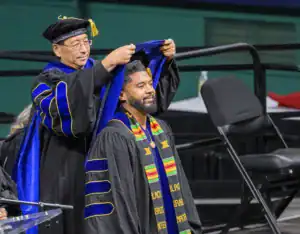 A graduate wearing a decorated gown and a colorful stole smiles as another person adjusts their hood during a graduation ceremony. A stadium setting with chairs is visible in the background.
