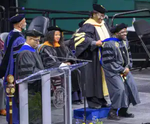 A graduate receives their hood during a commencement ceremony, while faculty members, including a woman adjusting the hood, and an audience of graduates and supporters look on. A podium is visible in the foreground.