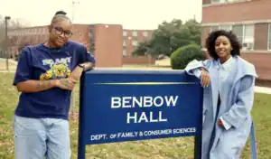 Two women stand beside a sign for Benbow Hall, Department of Family & Consumer Sciences. One wears glasses and a t-shirt, while the other is in a light blue coat, both smiling in front of the building.