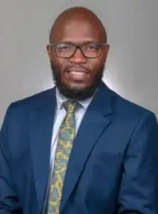 A man with glasses and a beard wears a blue suit and a patterned tie, smiling against a neutral background.