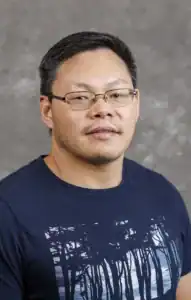 A man with short dark hair and glasses, wearing a navy blue shirt with a tree design, looks directly at the camera against a textured gray background.