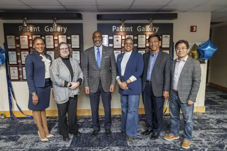 A group of six individuals stands in front of a wall displaying framed patents, celebrating a distinguished achievement in a professional setting. Colorful balloons are also present.