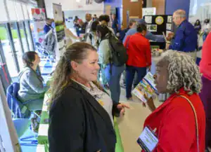 Two women engage in conversation at a bustling event. One wears a black blazer and gestures while the other holds a brochure, surrounded by people and informational displays.